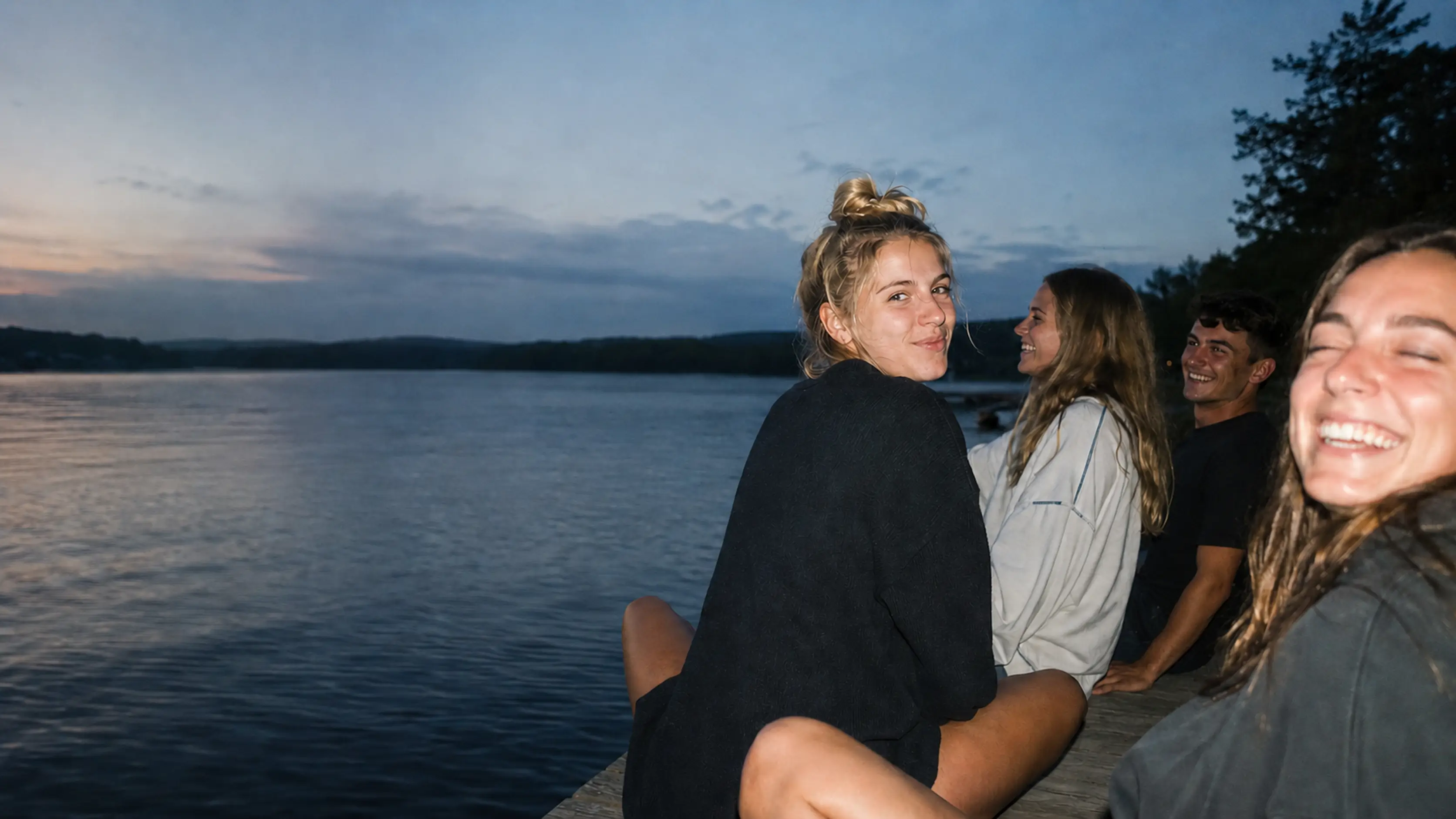 A group of friends sitting on a dock beside a lake at dusk.